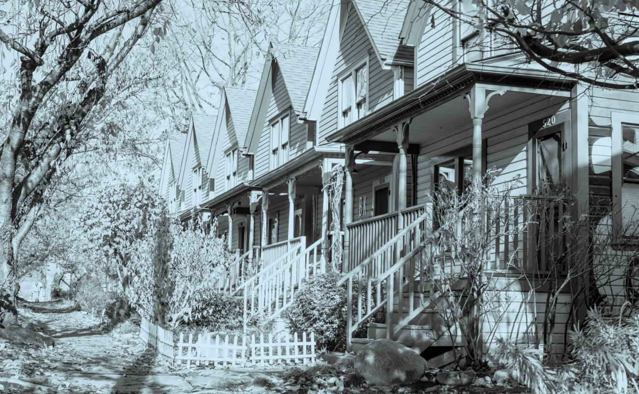 A row of identical, gabled townhouses with porches along a tree-lined street in the fall. The whole image has a blue overlay.